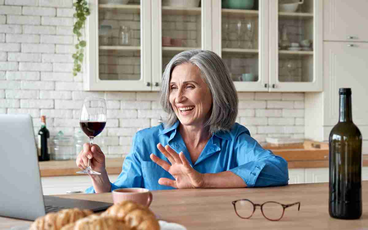 Happy 60s mature woman drinking wine video calling friend on laptop at home. Smiling old middle aged lady holding glass waving hand talking by online social distance chat meeting sit at kitchen table.